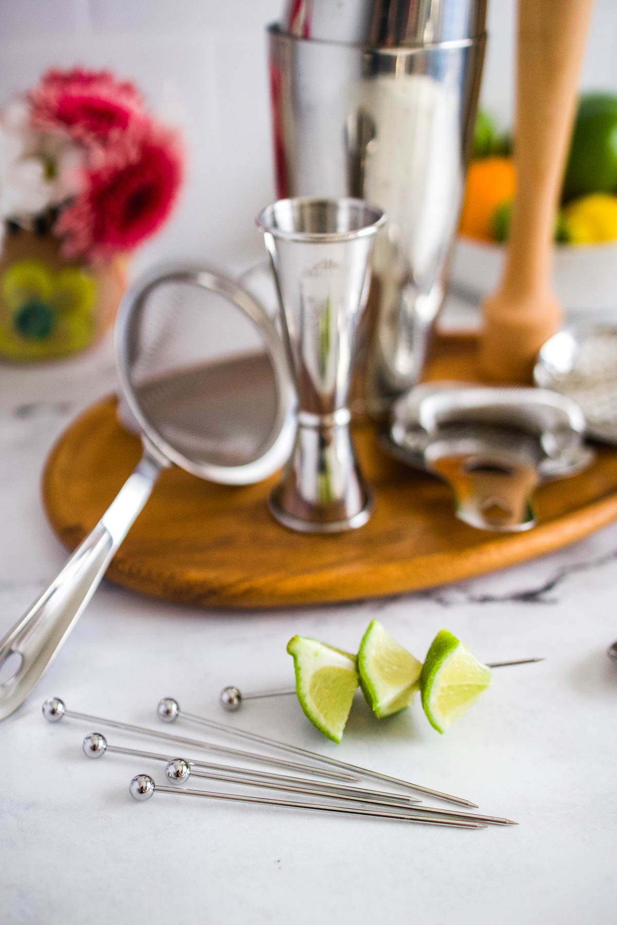 A fine mesh strainer next to stainless steel cocktail picks with lime chunks on them and a tray with other cocktail tools.