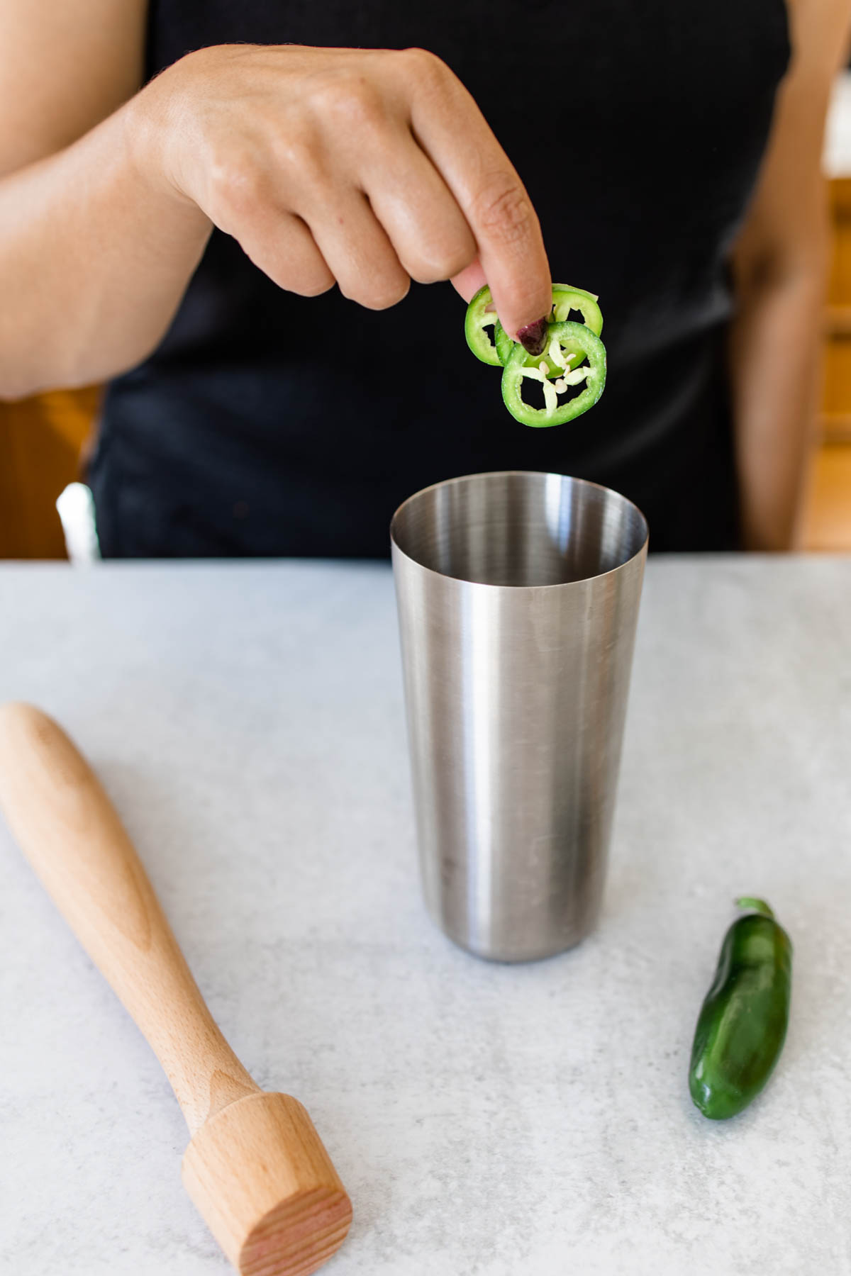 A cocktail shaker with a muddler on the table next to it and a woman adding slices of jalapeño into a cocktail shaker.