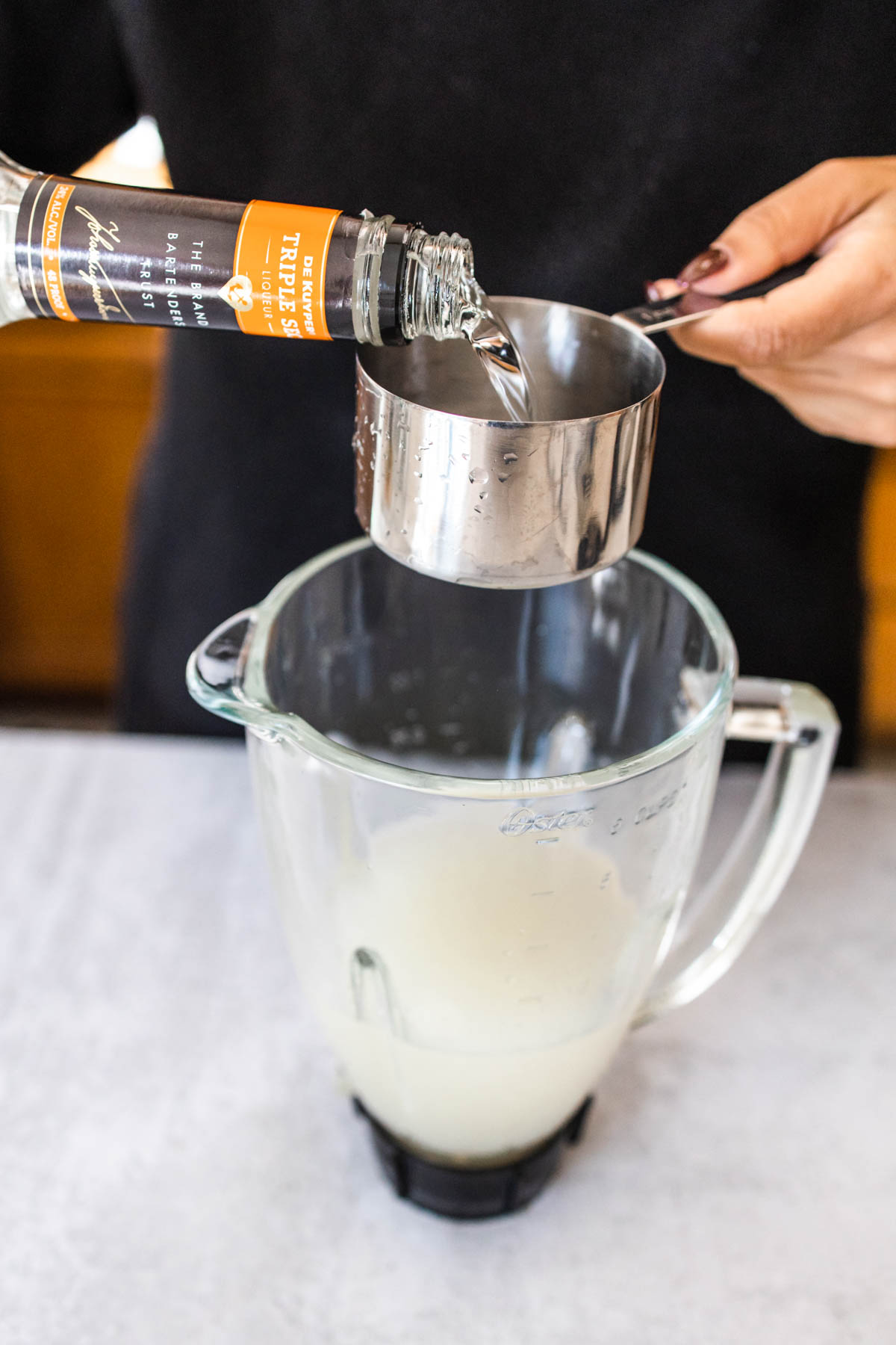 A woman using a measuring cup to add Triple Sec to a margarita pitcher.