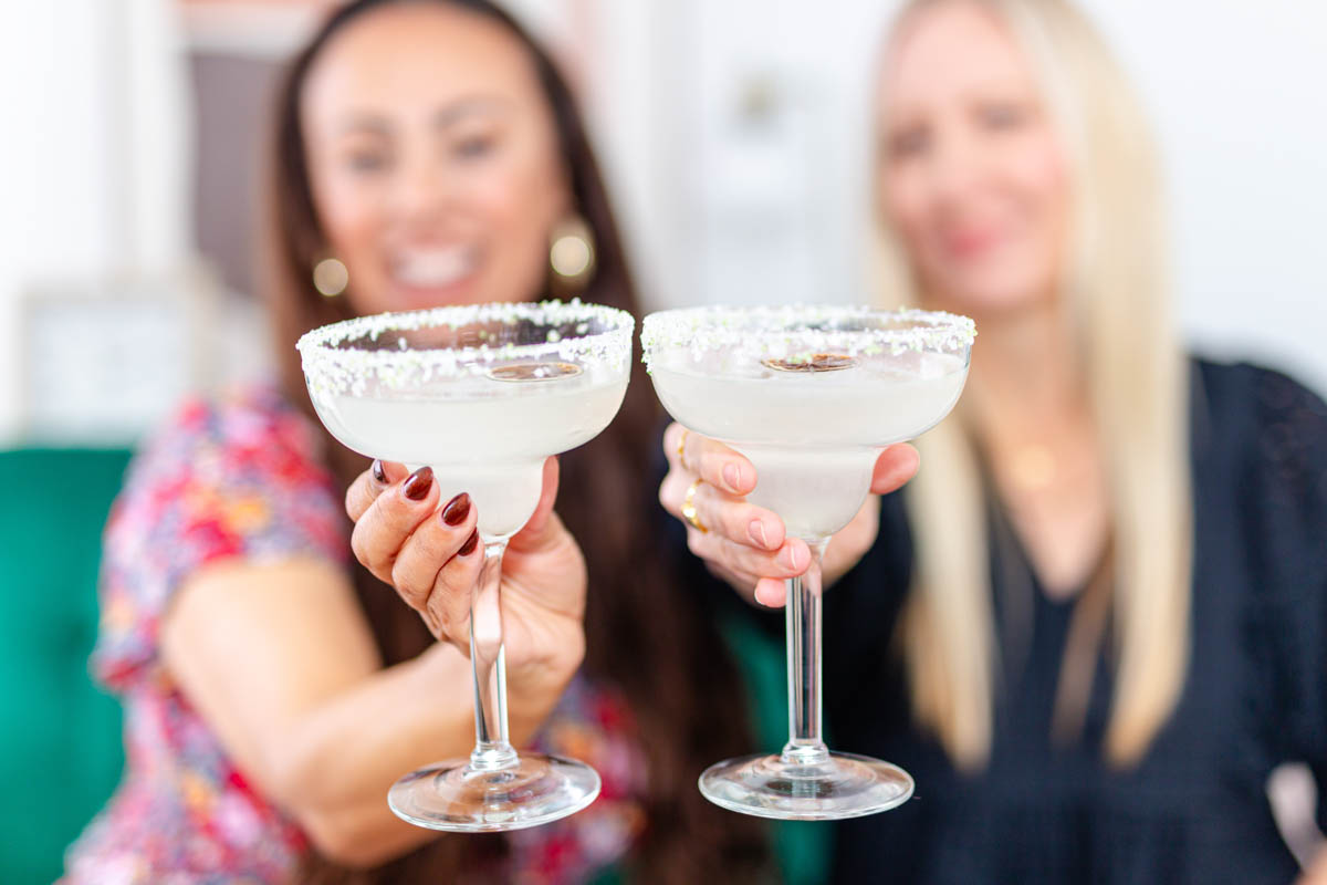 Two hands holding margarita glasses with women out of focus in the back.