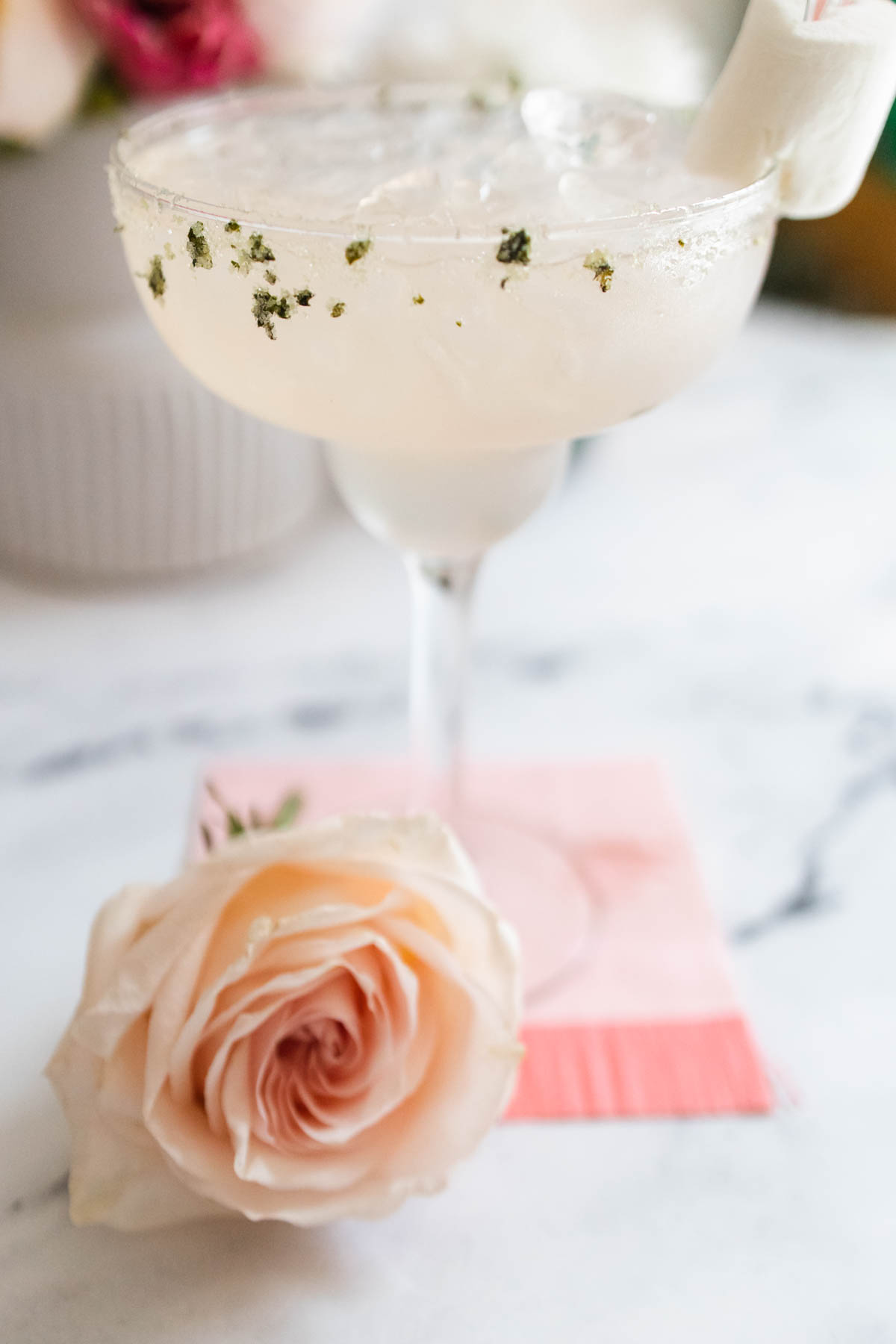 A close up of a margarita glass with a basil sugar rim on a table next to a pink rose.