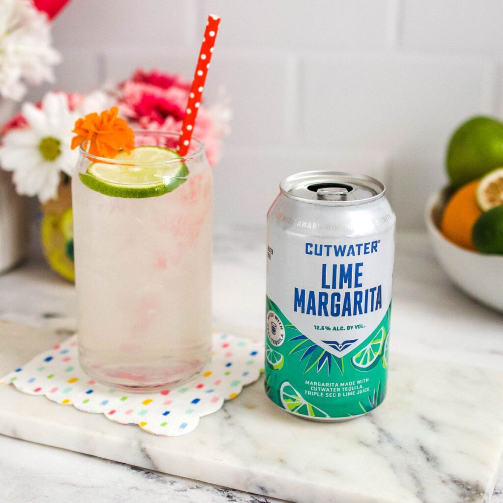 A can of Cutwater Lime Margarita on a table next to a glass with ice and a fun garnish.