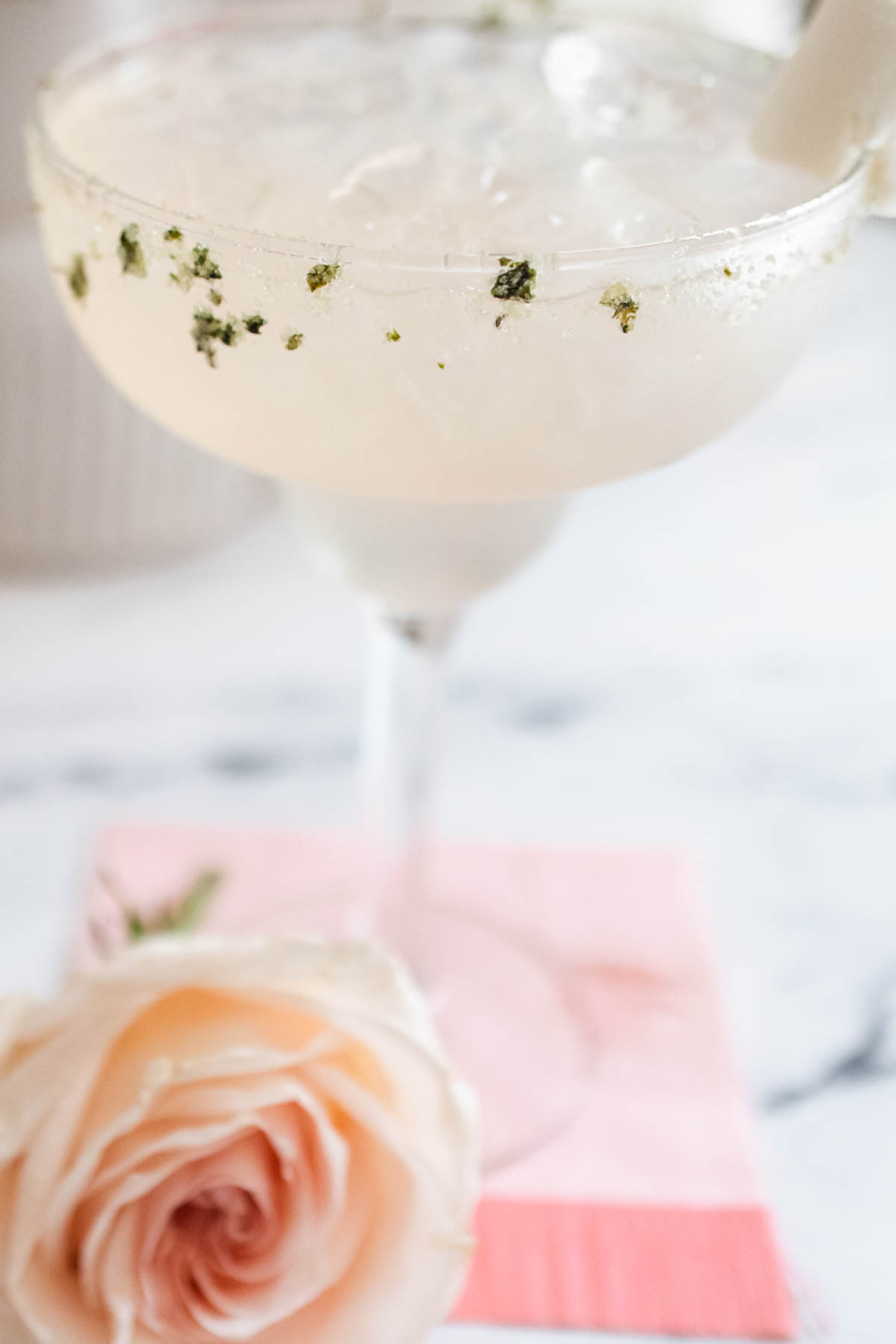 A margarita glass with a sugar rim with basil on a table next to a flower.