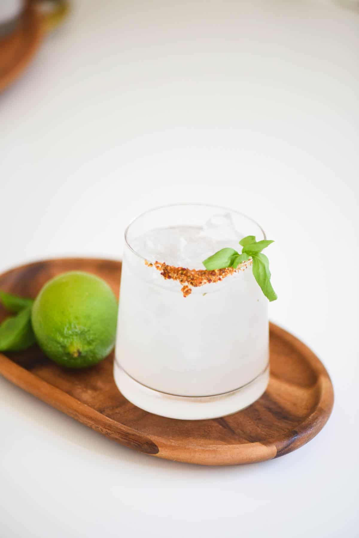 A pale white margarita served over ice in a glass with spice rim and fresh basil with a lime on the side placed on a wooden plate. 