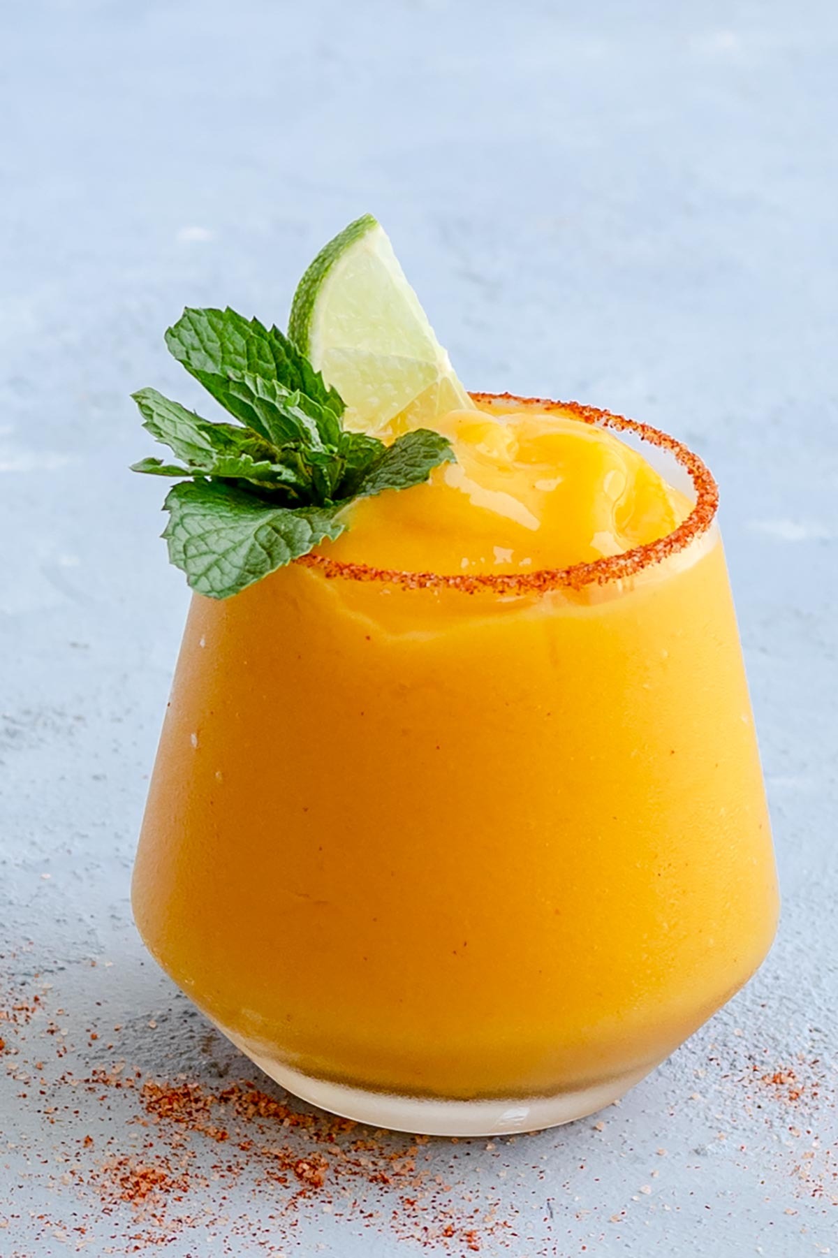 A yellow frozen mango margarita served in a glass with chili powder and sea salt rim as well as a lime wedge and mint sprigs. 
