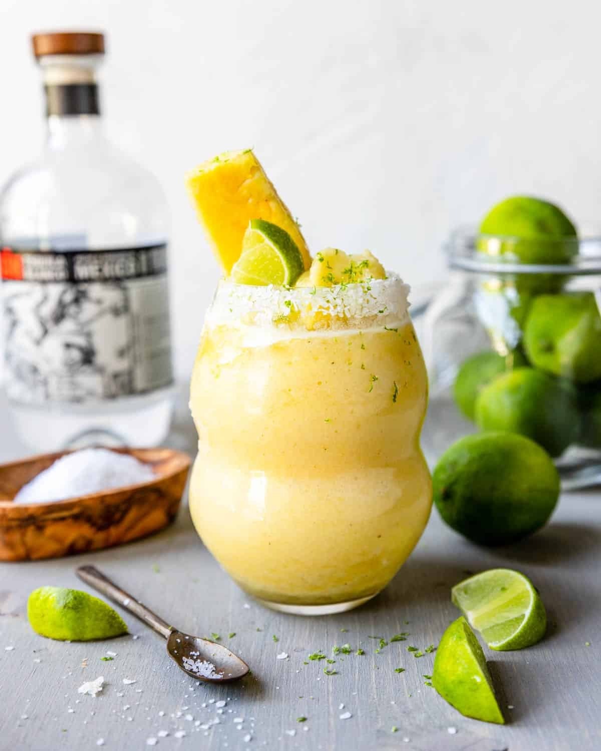 A yellow frozen pineapple margarita served with slices of pineapple and lime in a glass with sugar rim. 