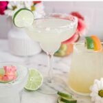 A margarita glass with a lime wedge on the side and a sugar rim, another glass with a slice of lime and pink flower petals on top, and a third smaller glass with a lime and orange slice.