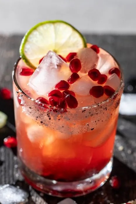 A pomegranate margarita served with pomegranate seeds, ice cubes, and lime wedge in a glass with salt and sugar rim. 