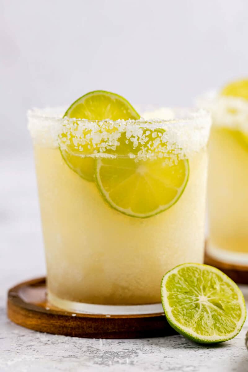 A pale yellow slushy margarita served with lime wedges in a glass with a salt rim.  