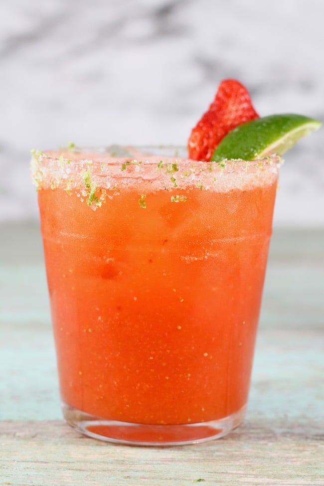 A vibrant pink margarita served with lime wedge and sliced strawberry in a glass with basil sugar rim. 