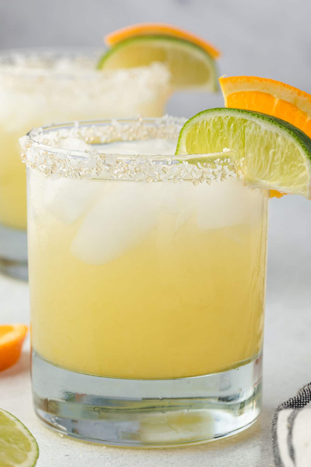 A pale yellow Texas margarita served with ice in a glass with salt rim and some lime and orange slices on the side. 