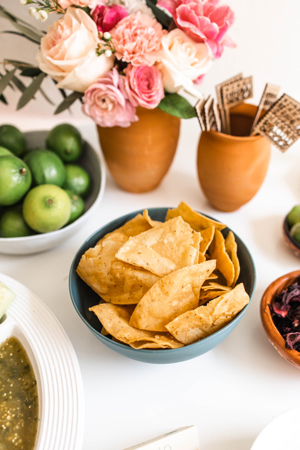 Tortilla chips in a small bowl on a party table next to some fresh flowers and limes.