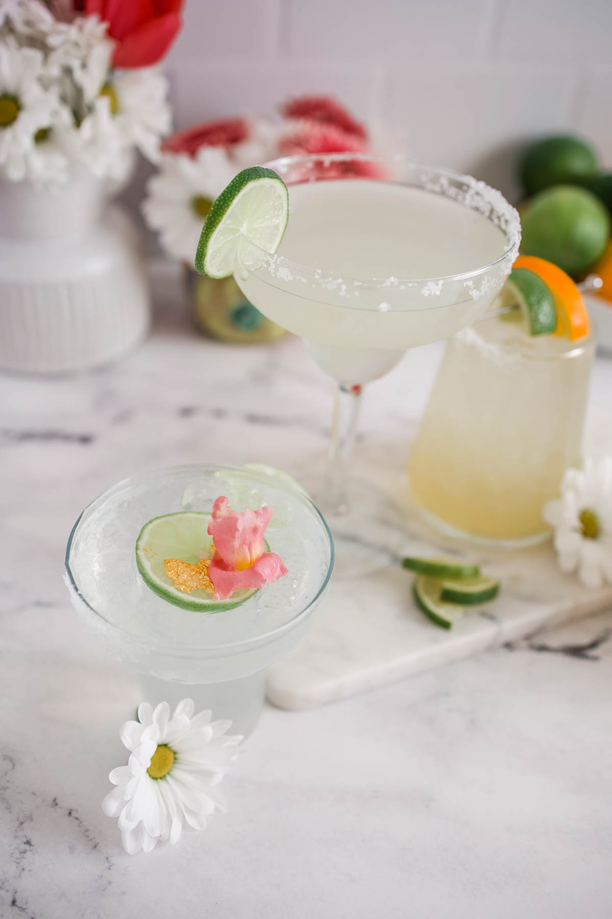 A margarita glass with a lime wedge on the side and a sugar rim, another glass with a slice of lime and pink flower petals on top.