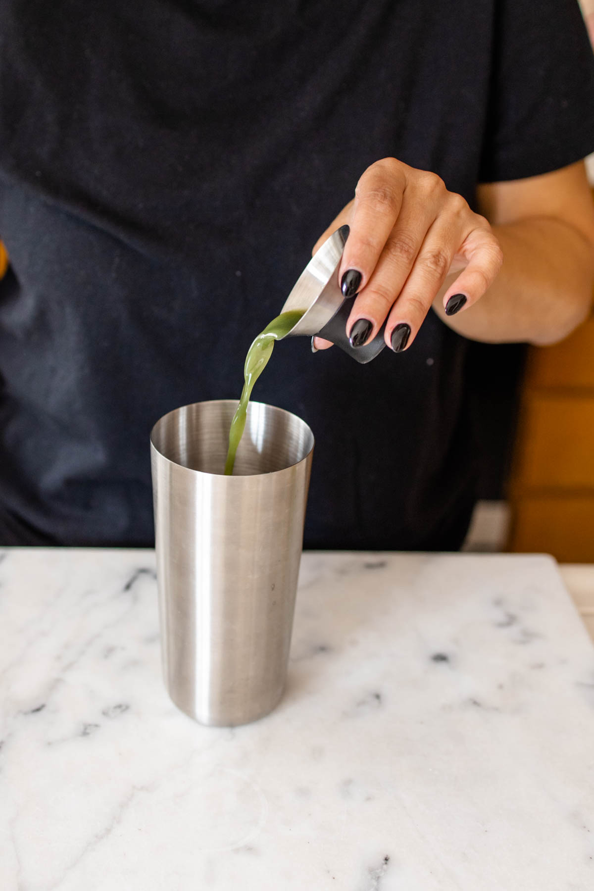 Woman adding green juice to a cocktail shaker from a measuring cup.