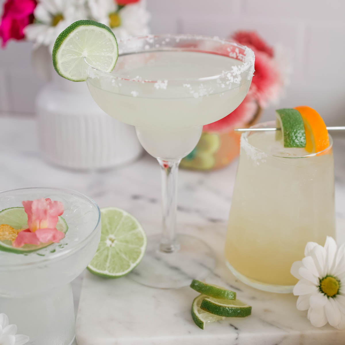 A margarita glass with a lime wedge on the side and a sugar rim, another glass with a slice of lime and pink flower petals on top, and a third smaller glass with a lime and orange slice.  