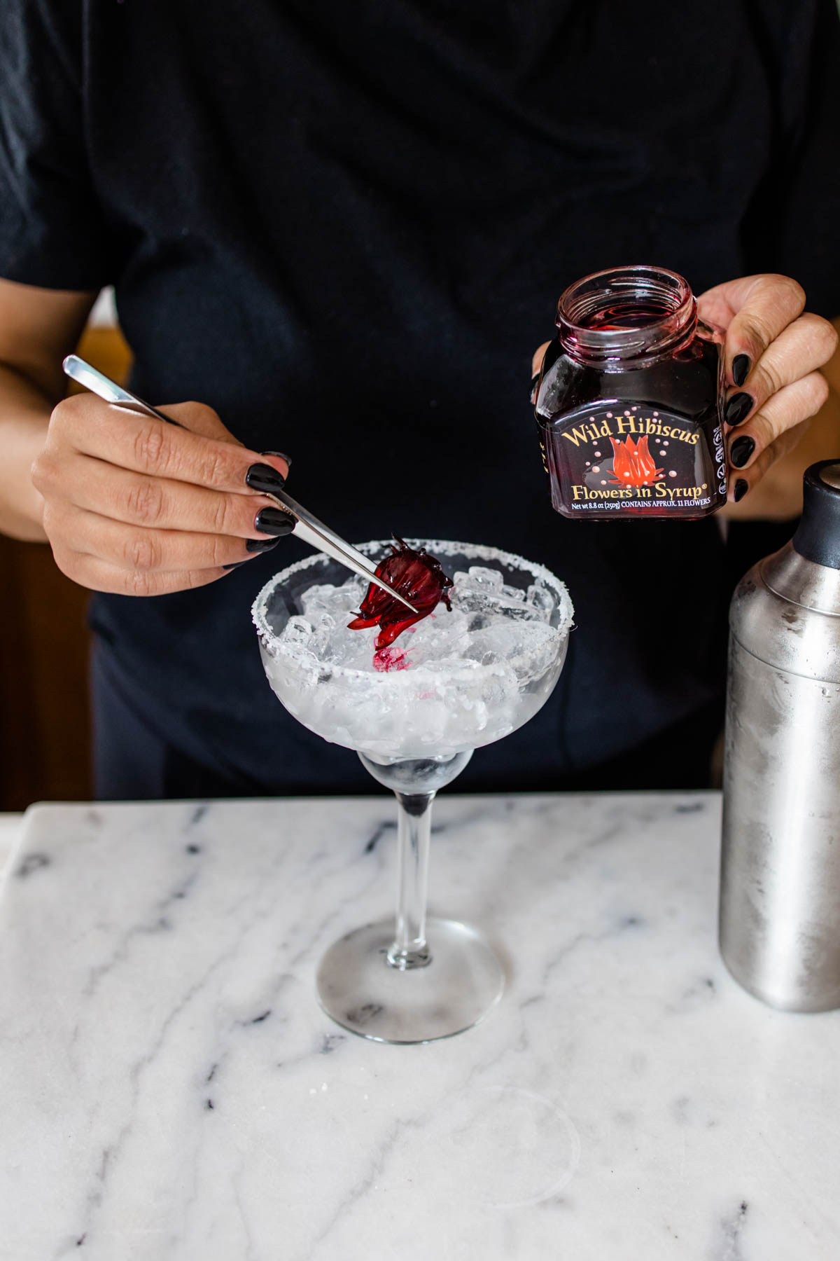 A salt-rimmed margarita glass with ice and hibiscus flower being placed on top of it. 
