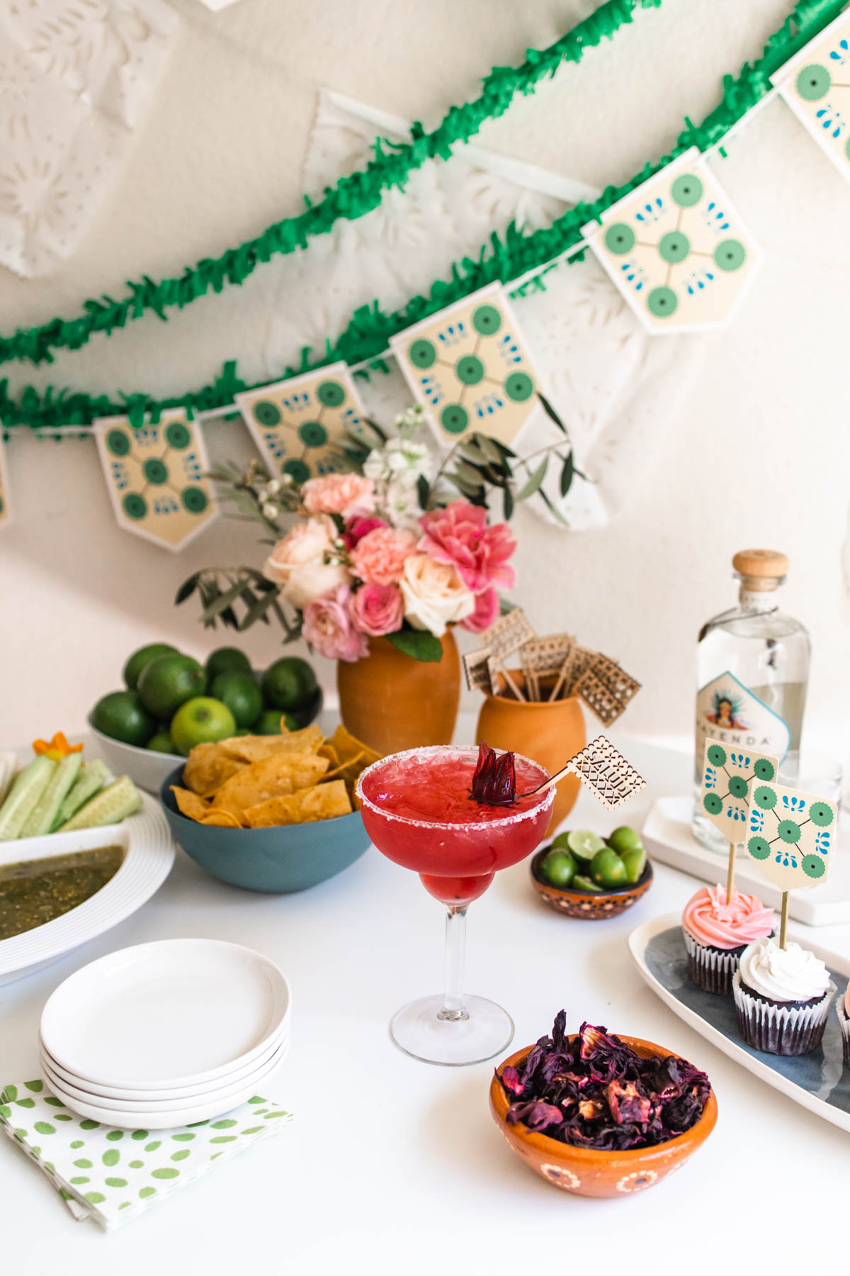 Hibiscus margarita in a salt-rimmed glass with hibiscus flower as a garnish with nachos, cucumber, lime wedges and cupcakes around it. 