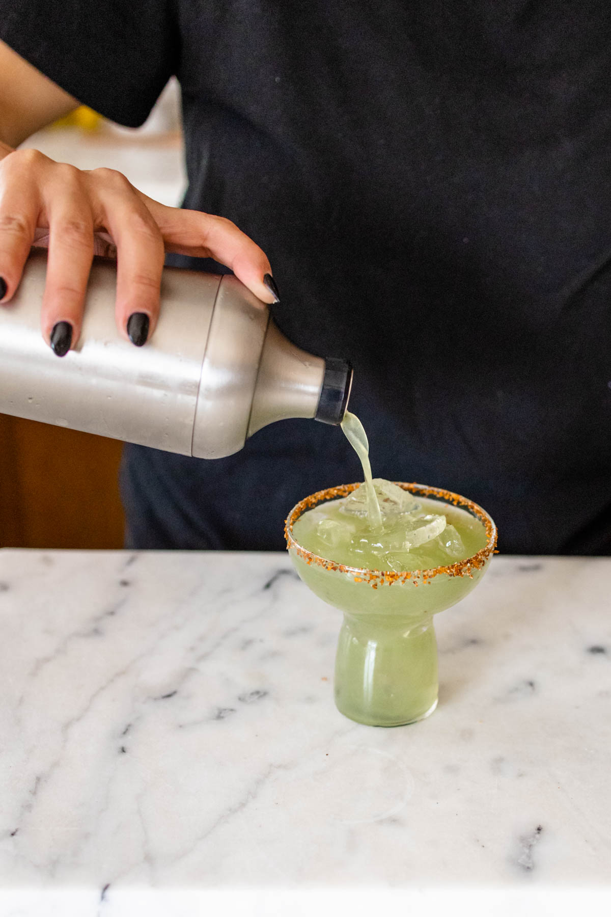 Woman pouring a green juice margarita into a stemless wine glass from a cocktail shaker.