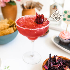 A margarita glass with salt rim and red hibiscus margarita with ice and hibiscus flower garnish.