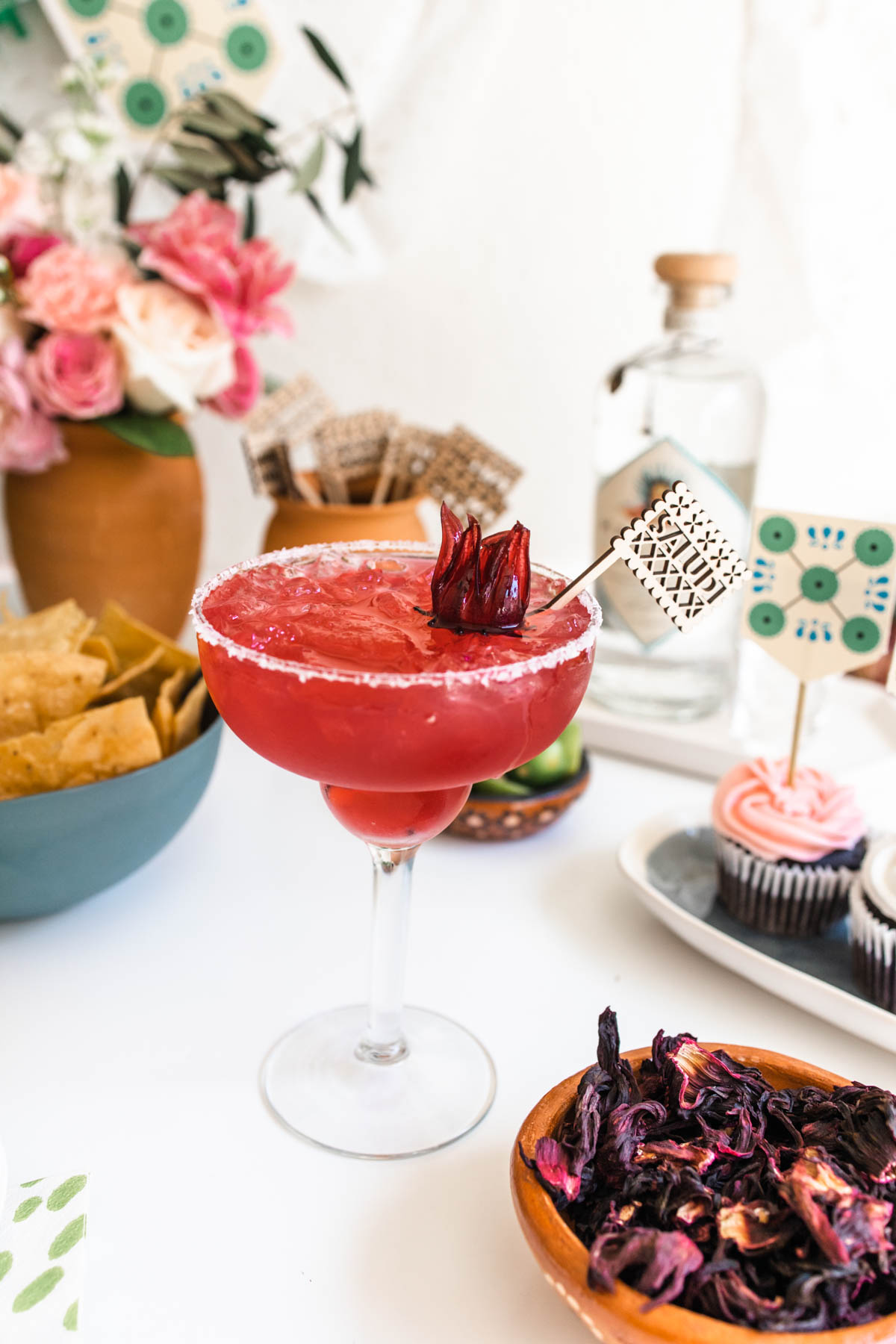 A gorgeous red hibiscus margarita is a salt-rimmed glass garnished with hibiscus flower and a tiny flag with "Salud" written on it. 