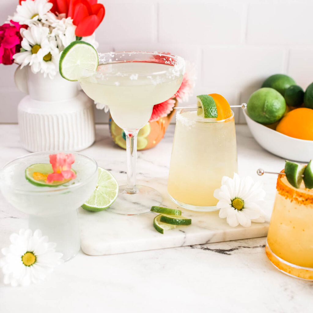 A collection of margaritas on a table with cut limes and daisies.