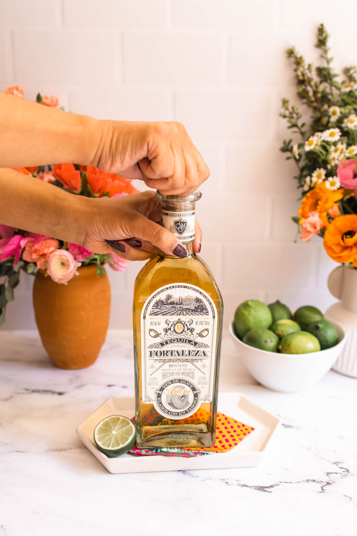 A hand opening a bottle of Fortaleza Anejo Tequila that's beside a half slice of lime on a white hexagon plate.