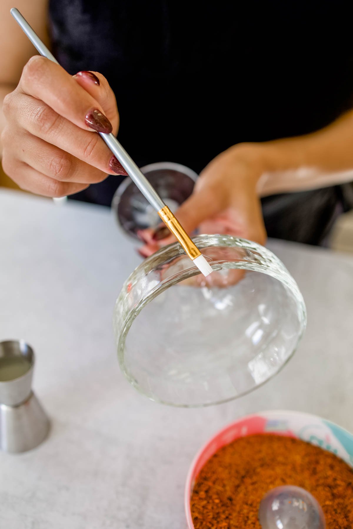 A woman moistening a portion of the rim of a margarita glass using a thin brush.