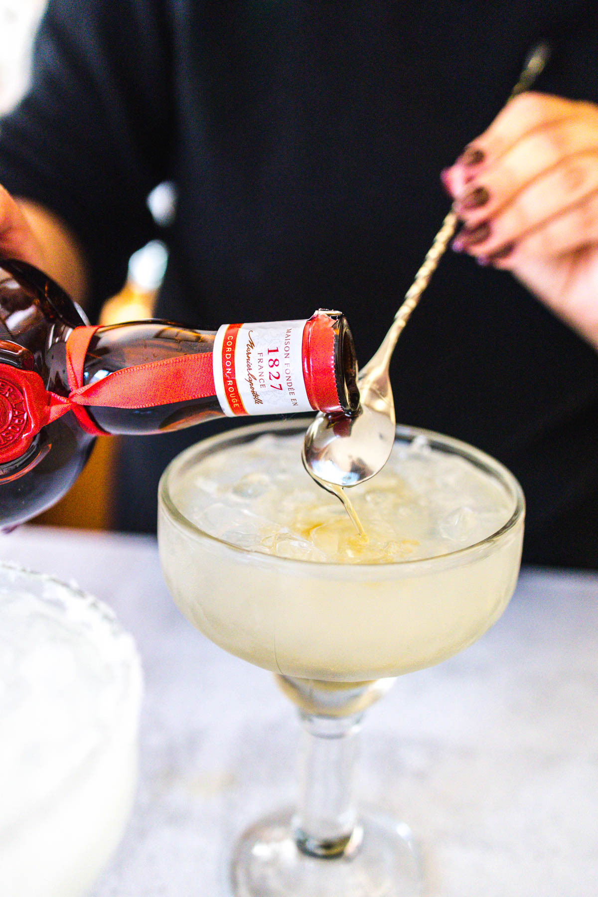 A woman pouring some Grand Marnier over the back of a bar spoon on top of a margarita in a glass.