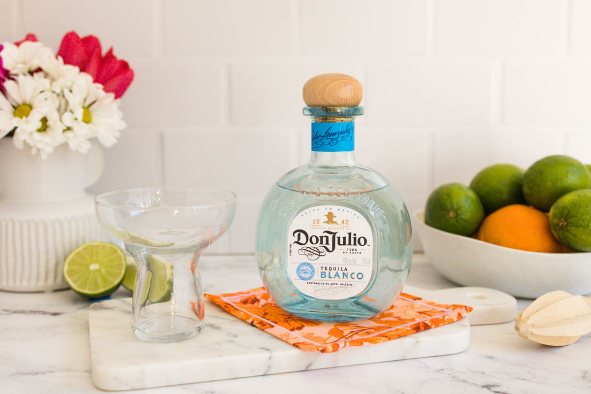 Bluish decanter of Don Julio tequila blanco with a wooden top beside a stemless margarita glass on a counter.