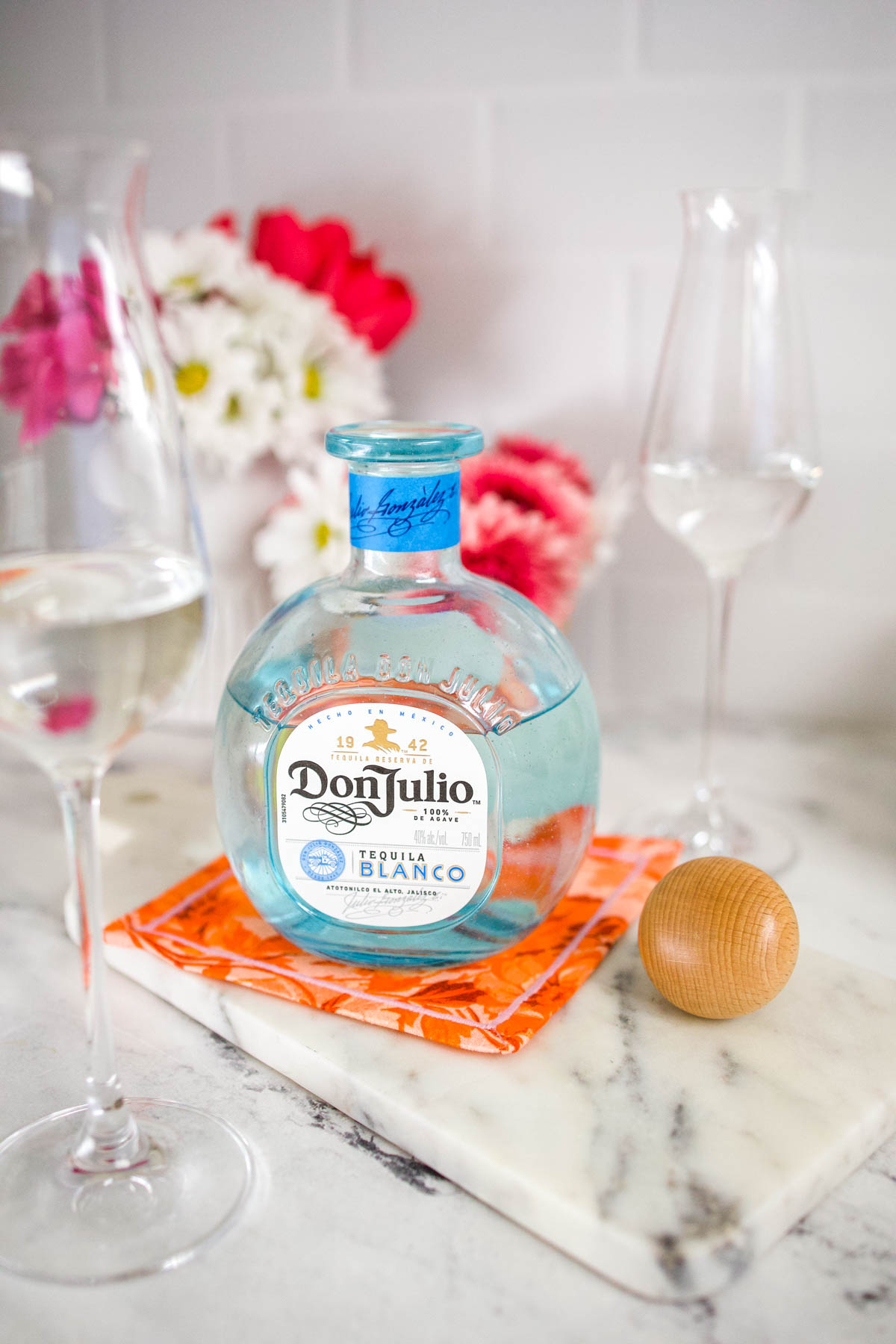 A bottle of Don Julio Blanco Tequila in between two tall flute glasses and a wooden cork on a counter.