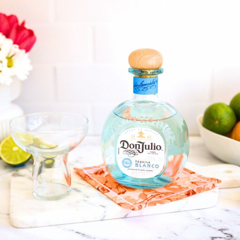 A stemless margarita glass alongside a beautiful light blue bottle of Don Julio Blanco tequila on top of an orange piece of cloth.