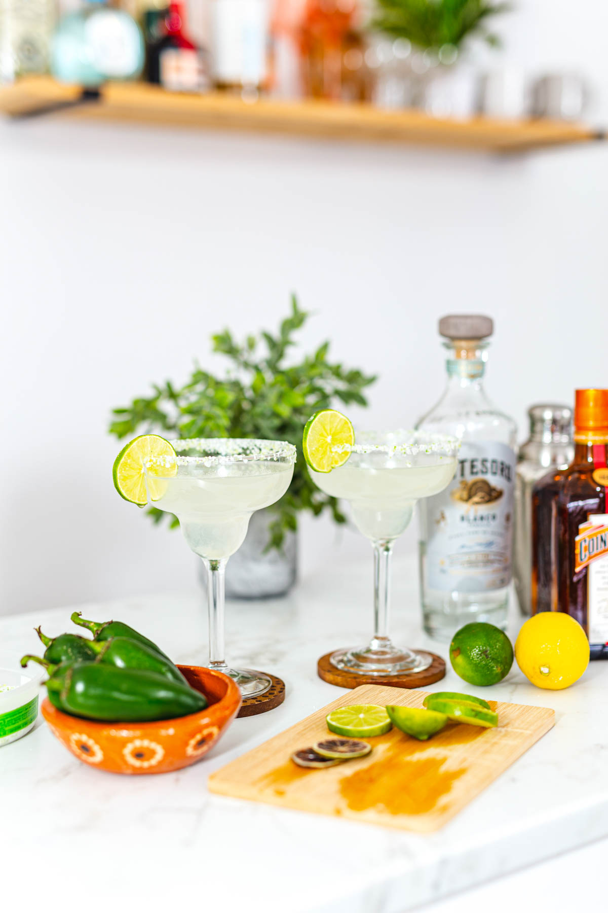 Two margarita glasses alongside jalapeno peppers, salt, lime, lemon, and bottles of El Tesoro Blanco Tequila and Cointreau.