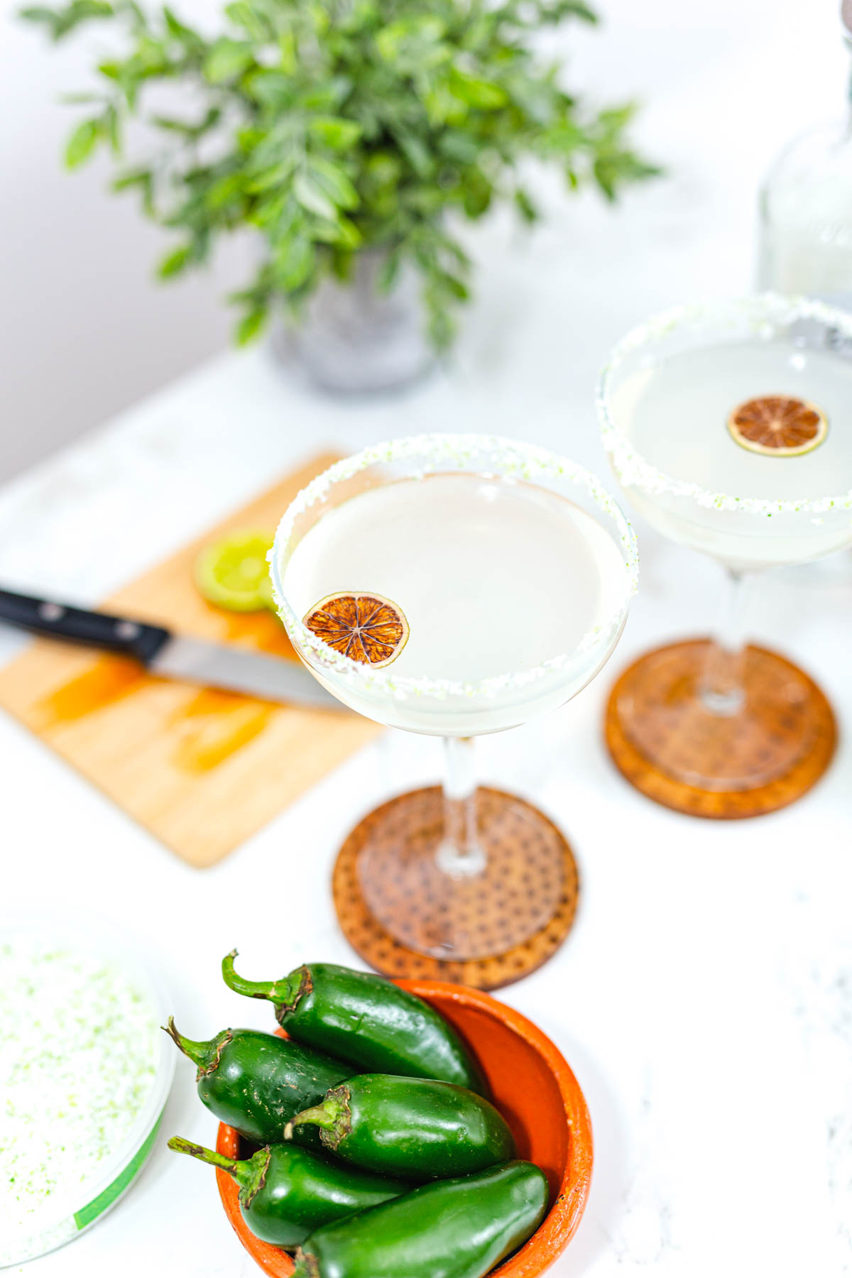 Two salt rimmed margarita glasses with a slice of dried lime as garnish on a table next to a bowl of jalapenos.
