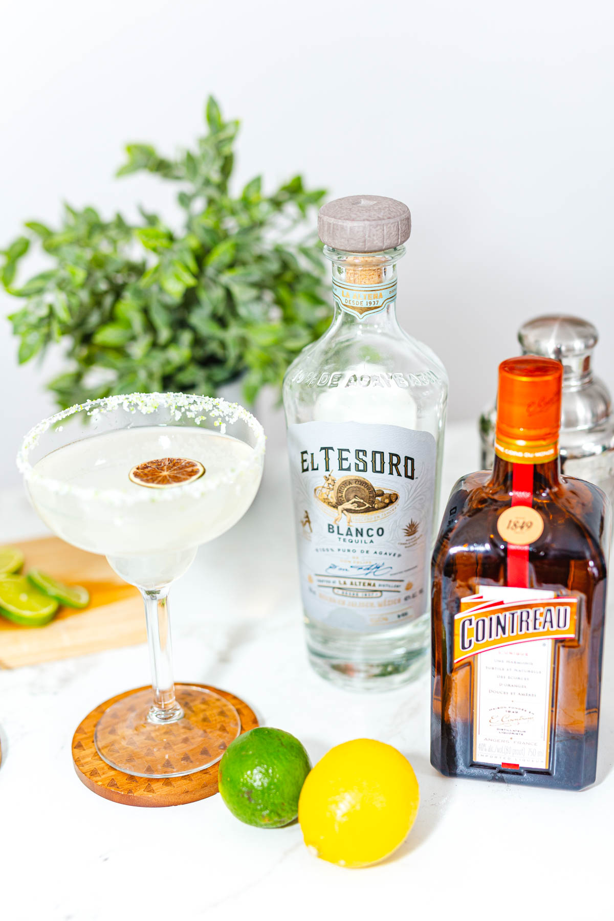A margarita glass alongside lime, lemon, and bottles of El Tesoro Blanco Tequila and Cointreau.