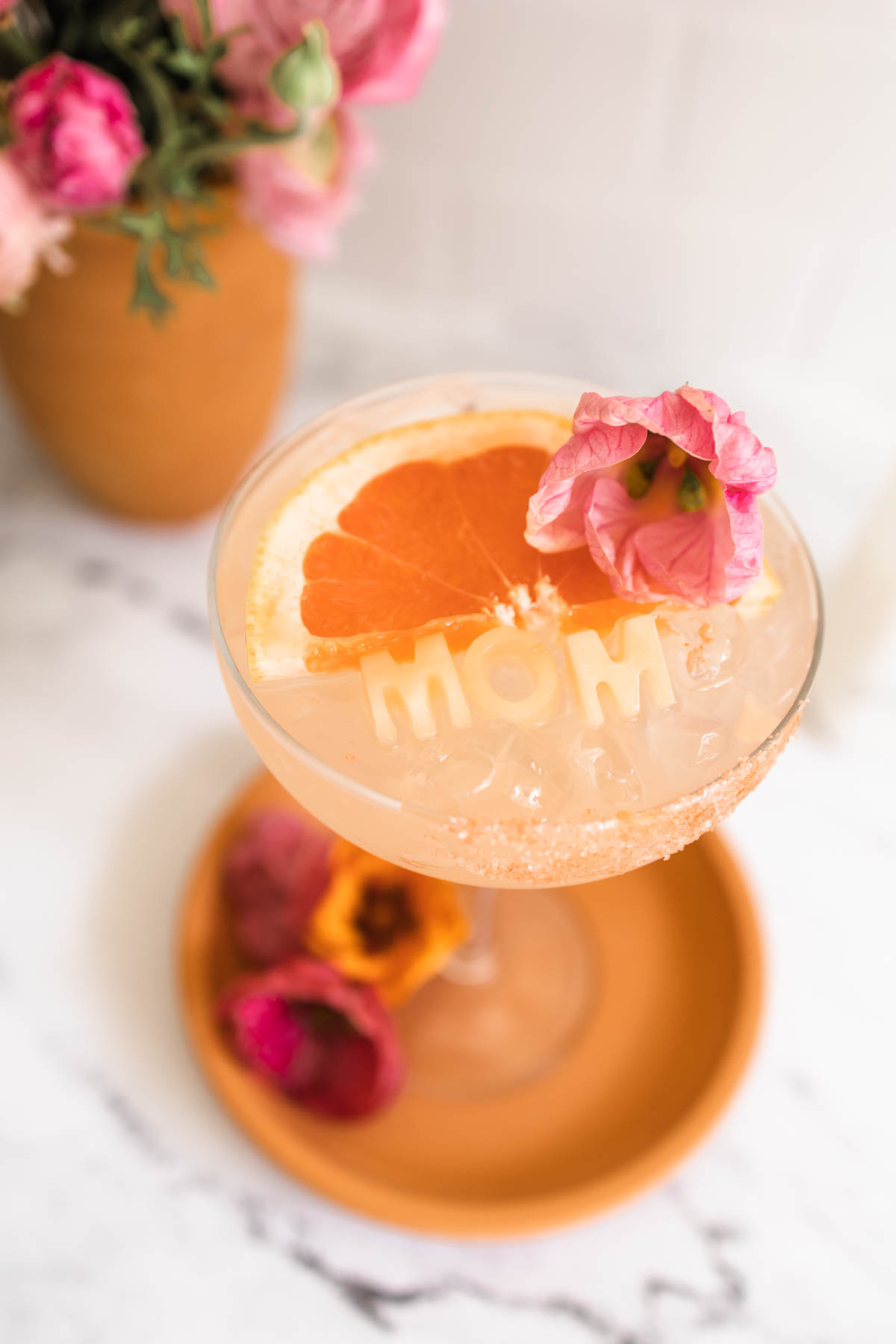 A rosé margarita in a margarita glass garnished with grapefruit wedge, a pink flower, and grapefruit-flavor salt rim as well as ice cubes that say "mom".