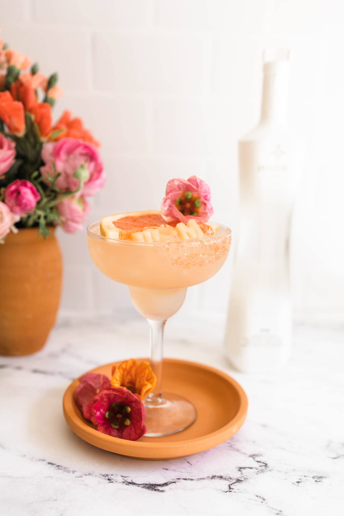 A lovely pink rose margarita on a margarita glass garnished with grapefruit wedge, a pink flower, and grapefruit-flavor salt as well as MOM ice cubes.
