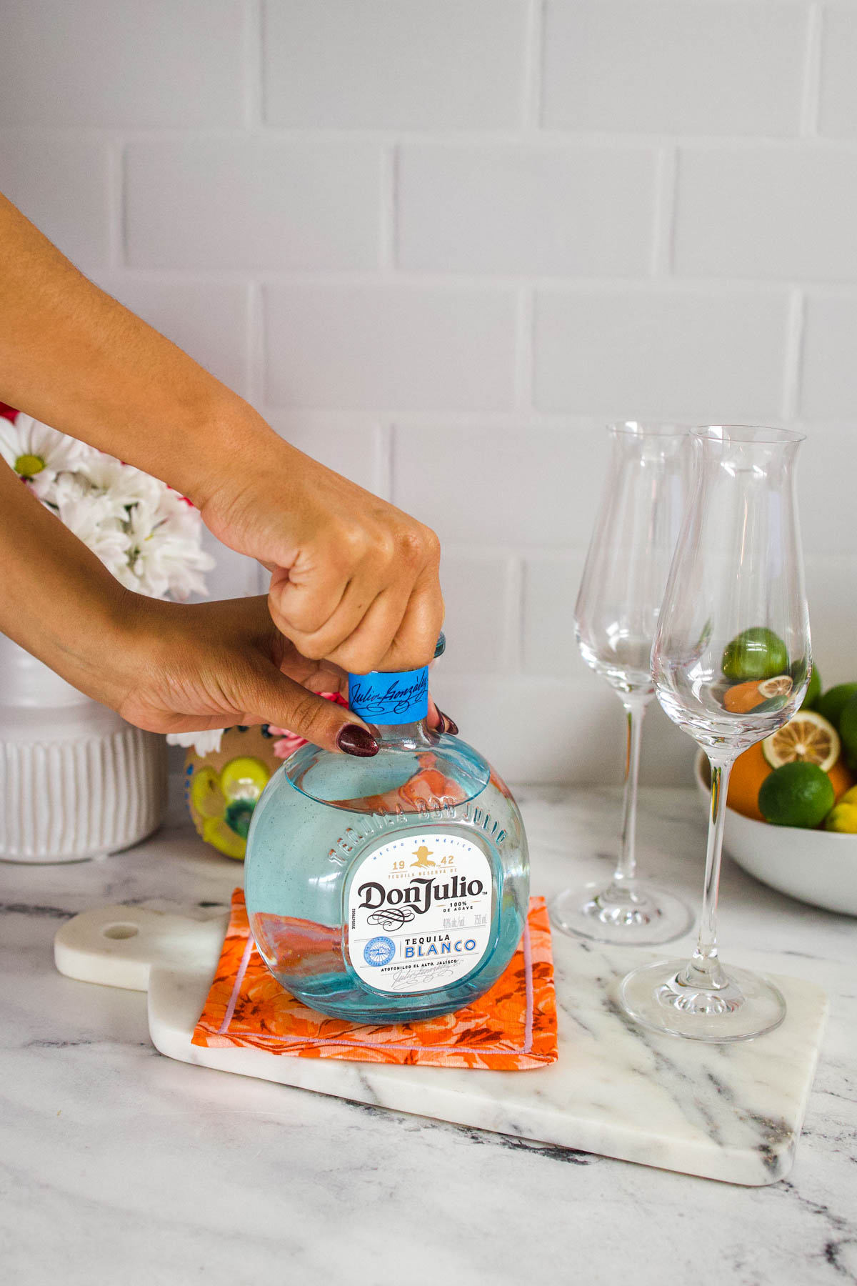 A hand opening a bottle of Don Julio Blanco Tequila right beside two flute glasses on a marble countertop.