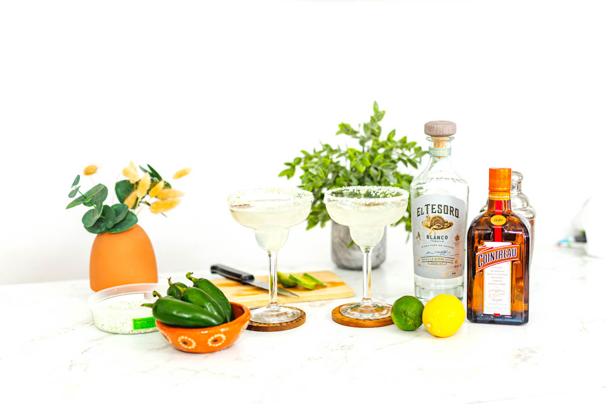 Two margarita glasses on a table with ingredients to make the drink including a bottle of tequila, triple sec and a lime with other decorative touches around it.