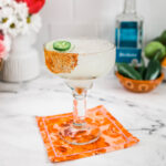 A margarita glass with Tajin rim filled with spicy margarita on an orange napkin.
