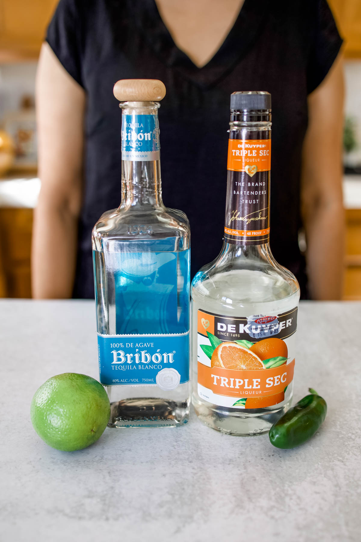 Bottles of Tequila Blanco and Triple Sec along with a fresh lime and jalapeno on a counter.