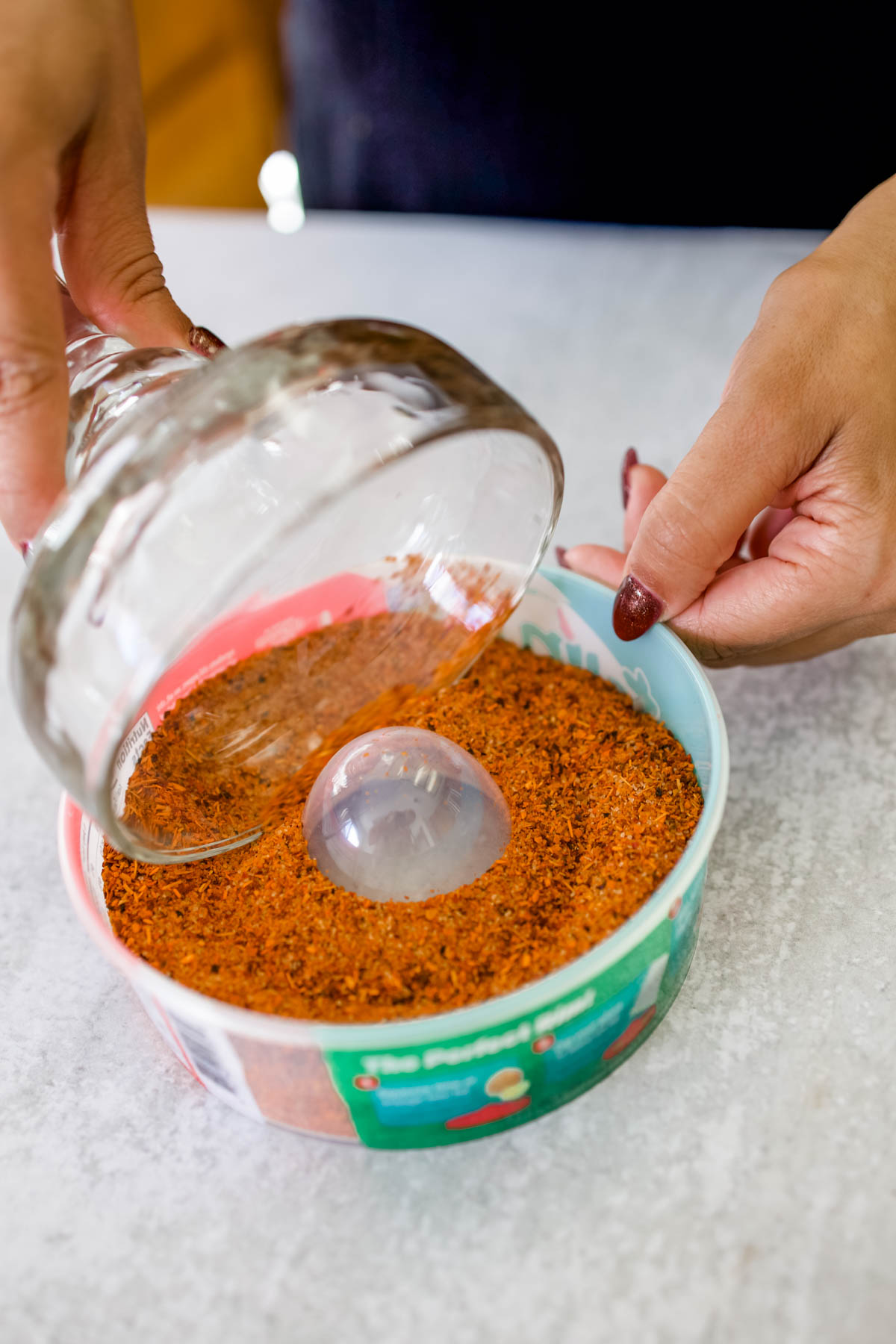 A hand dipping one side of the margarita glass in a Tajin and salt mixture on a small plastic container.