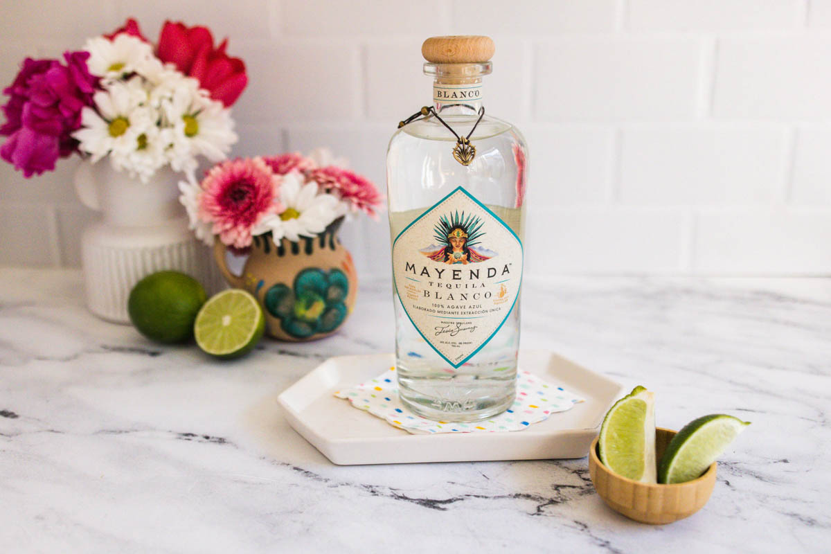A bottle of Mayenda blanco tequila on a table next to cut limes with decorative flowers in the background.