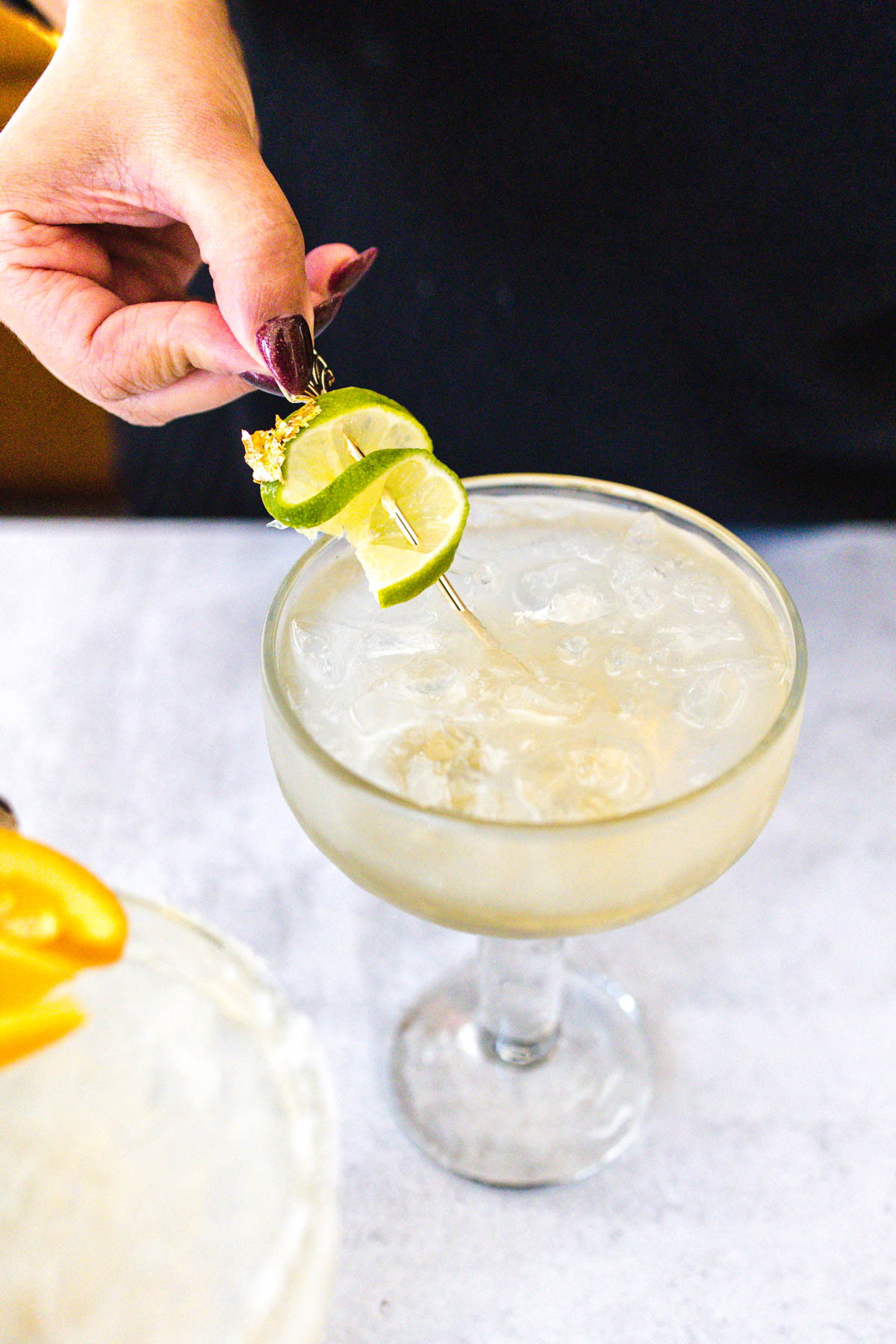 A Cadillac margarita in a glass with a woman holding skewered lime wedges on a stick.