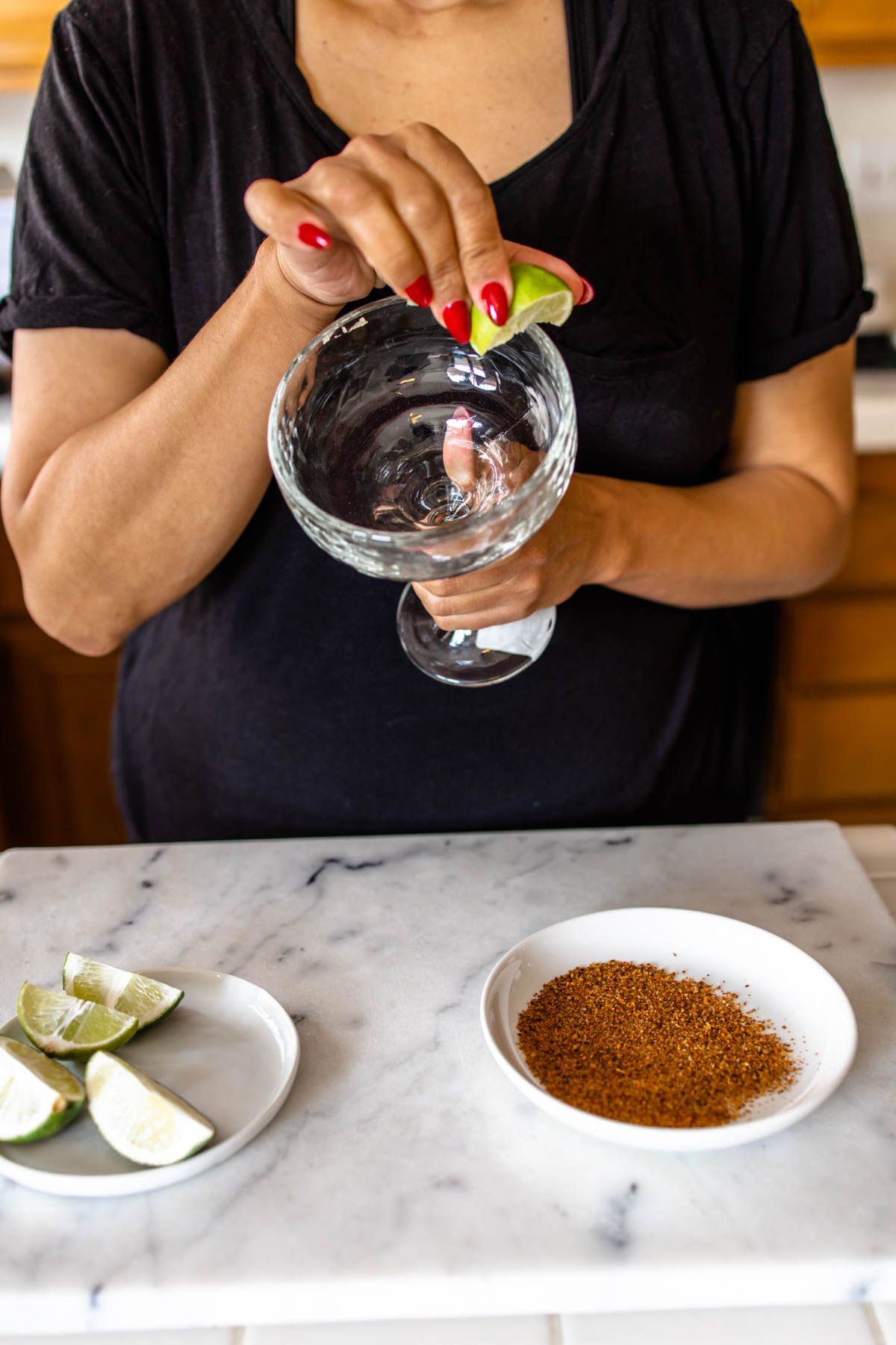 A woman moistening the rim of a margarita glass with a lime wedge with a small plate of lime wedges and tajim salt on the marble countertop.
