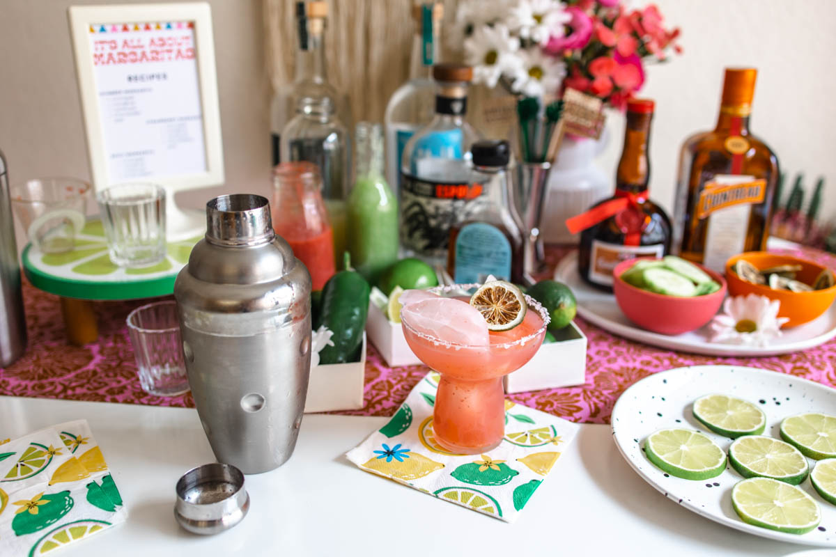 A cocktail shaker beside a strawberry margarita in a salted rim glass garnished with a dried lime wheel, as well as a plate of sliced limes, in front of various margarita ingredients.
