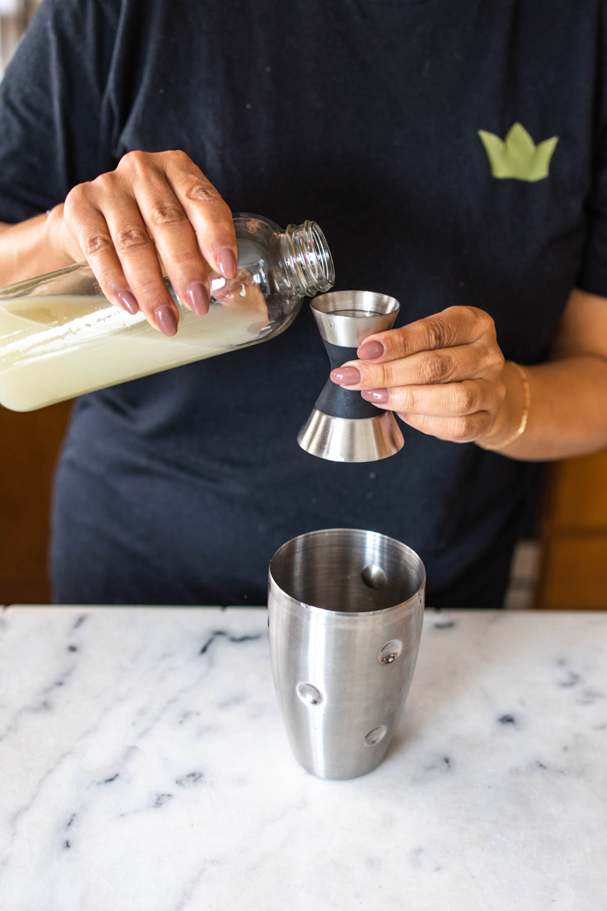 A person pouring some pineapple juice into a jigger behind a cocktail shaker on a marbled counter.