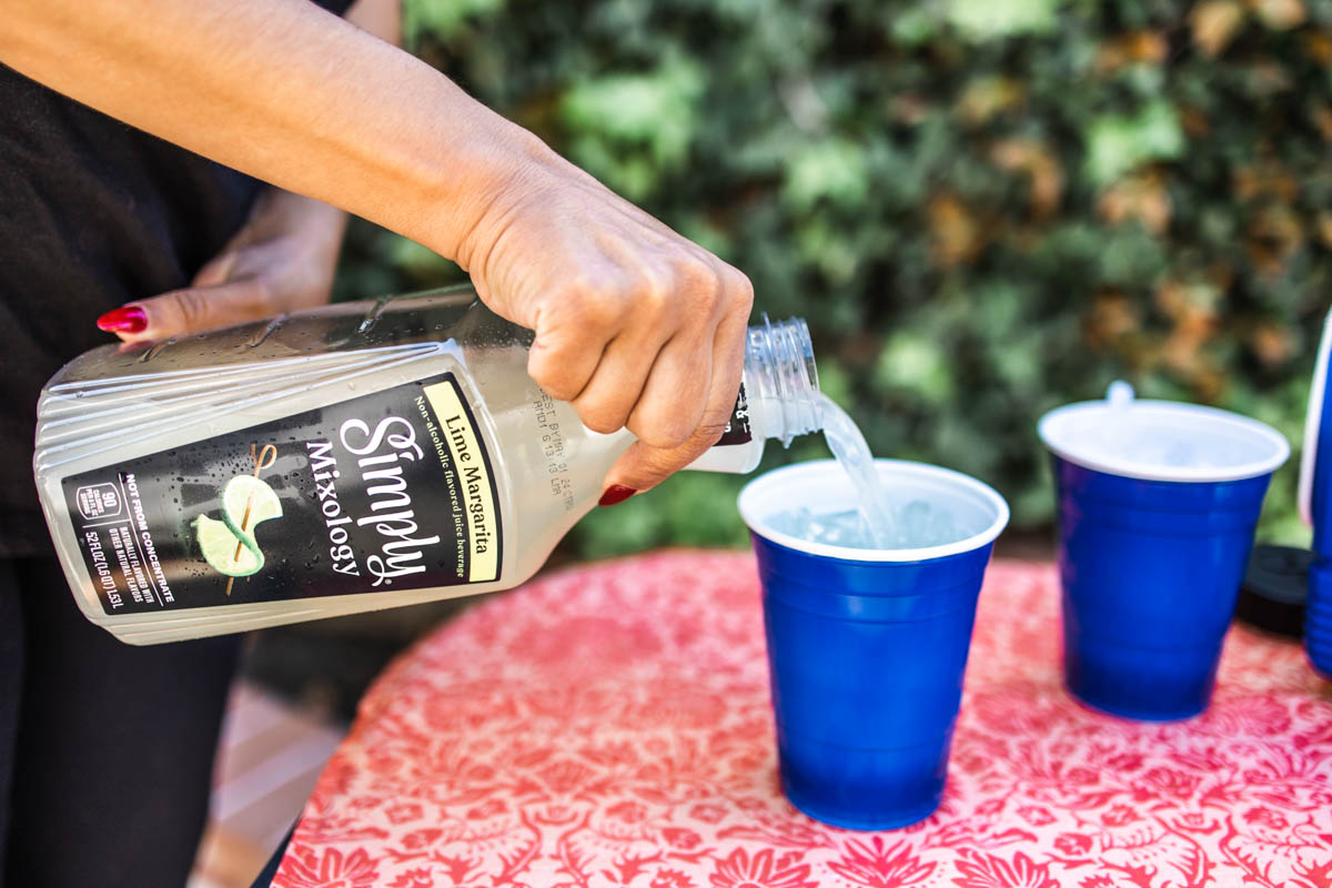 A person pouring to go margaritas from a Simply Mixology bottle into one of two blue plastic cups.