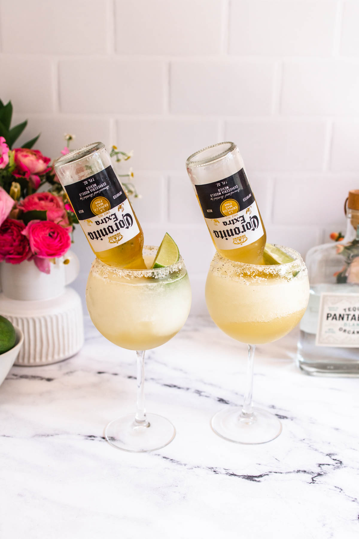 Two large margarita glasses with frozen beergaritas inside garnished with inverted Corona Light beers. 
