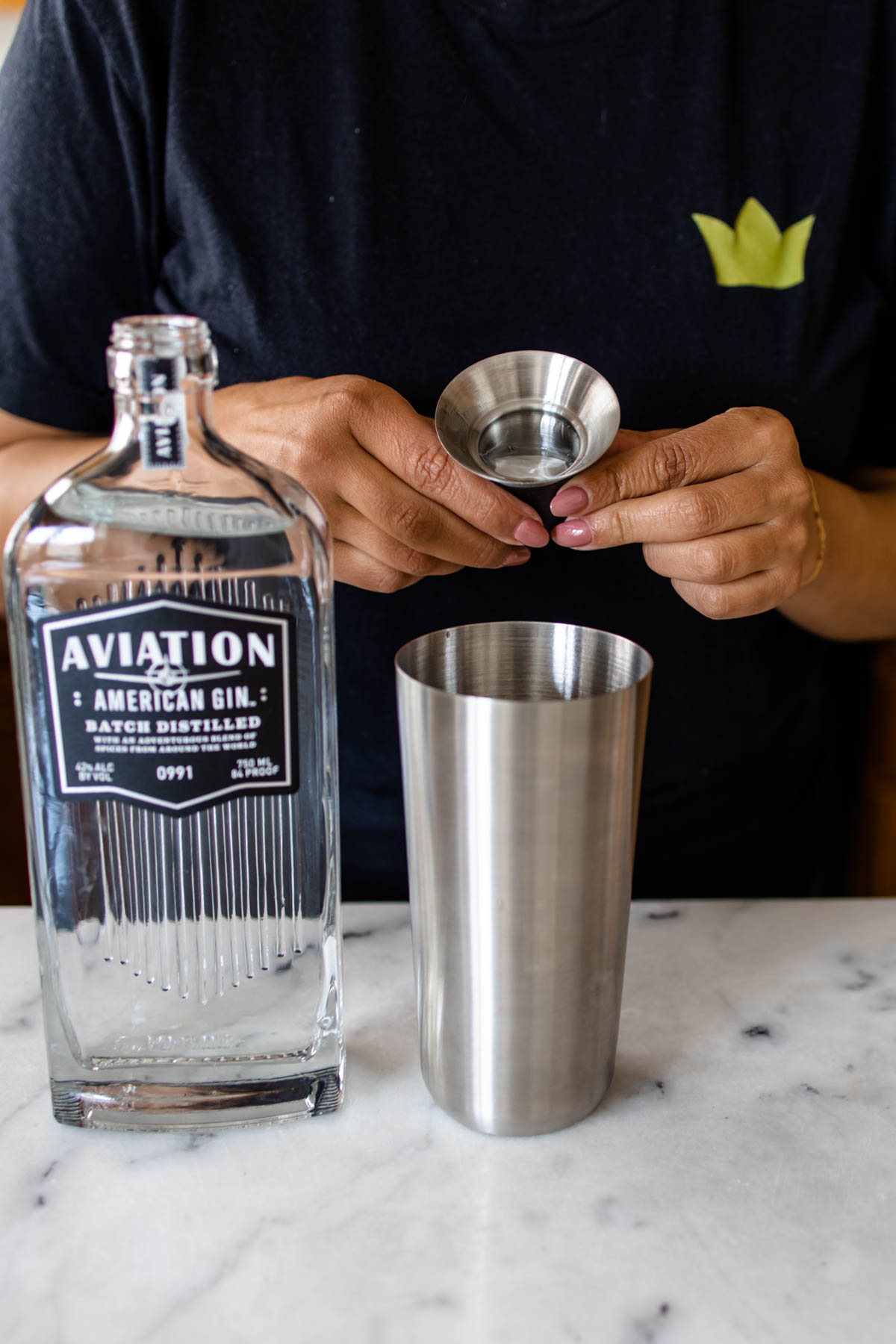 A person pouring some Aviation Gin into a cocktail shaker from a jigger, with a bottle of Aviation Gin beside it.