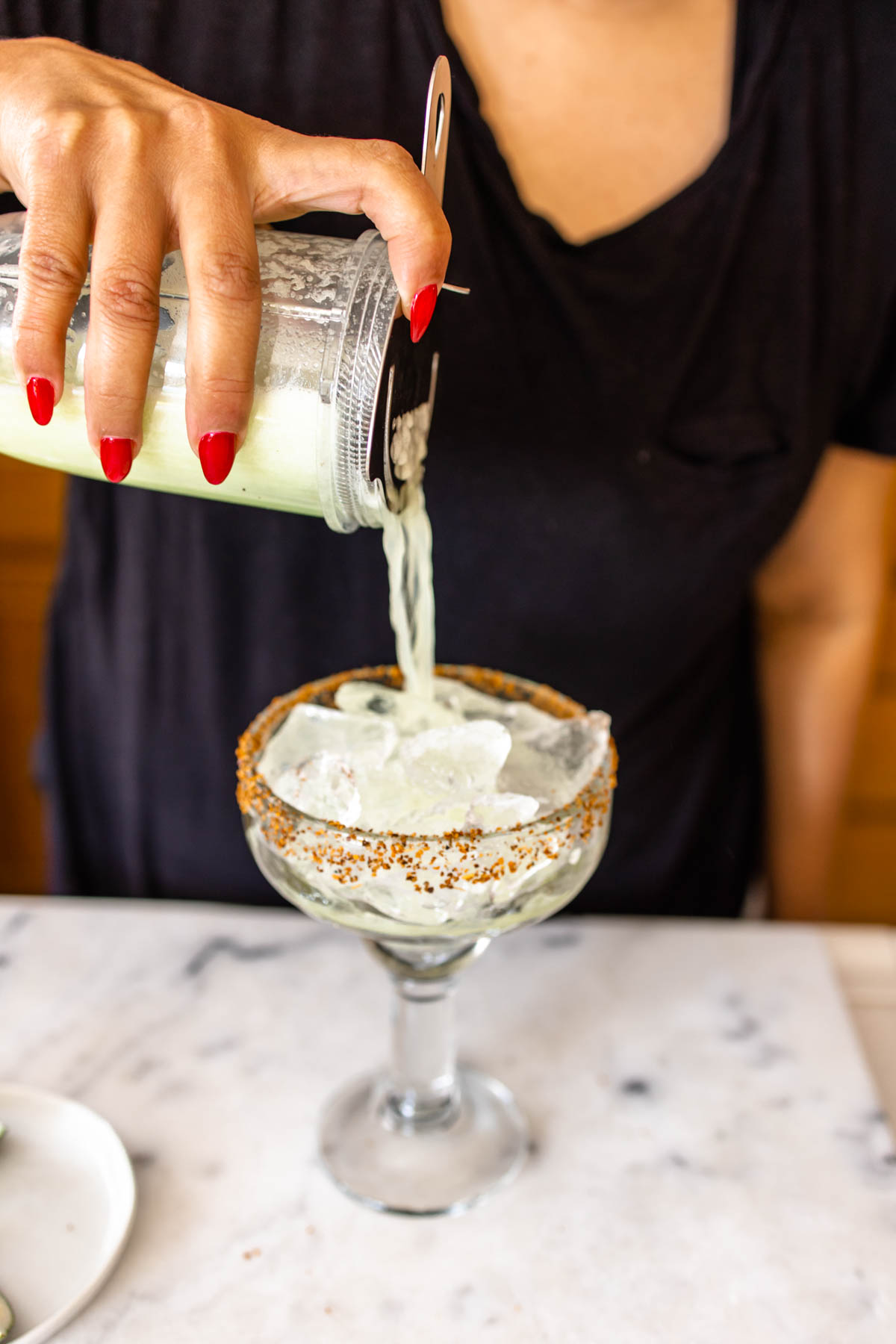 A woman straining the margarita mixture from a blender to the glass with Tajin rim using a Hawthorne strainer.